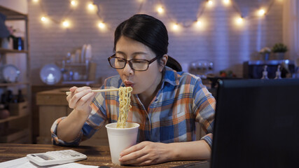 lazy smiling young asian chinese woman eating instant noodles by chopsticks in kitchen at home in evening. elegant girl in eyeglasses mouth blowing ramen soup while overtime work on laptop computer.