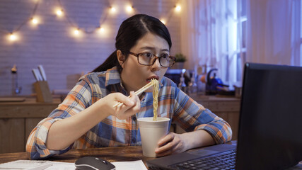 young japanese girl eats instant noodles and looks in laptop computer monitor. serious starving...