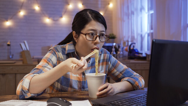 Young Japanese Girl Eats Instant Noodles And Looks In Laptop Computer Monitor. Serious Starving Office Lady Worker Working At Home In Night Kitchen And Having Bedtime Snack With Unhealthy Fast Food