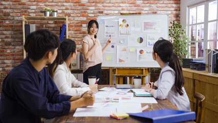 smiling asian japanese businesswoman presenting to colleagues at meeting in boardroom. charming girl employee holding marker point on whiteboard with charts showing coworkers report of new project.