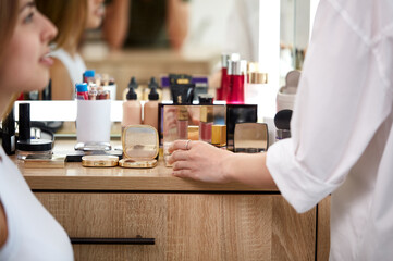 close-up photo of professional visagist's hand taking decorative cosmetics from table, going to apply powder on model's face. in beauty studio