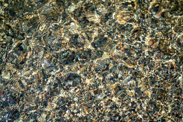 The texture of the bottom. Many small stones are visible under the clear water. Water ripples of the sea, sun rays on the rocks.