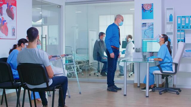 Senior man with face mask against covid-19 at hospital reception talking with nurse. Doctor with patient in examination room. Disabled mature woman with walking frame in waiting area.