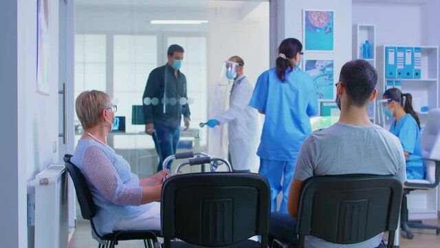 Private hospital reception area during global pandemic. Doctor with visor against coronavirus inviting man in his office for examination. Disabled senior woman with walking frame