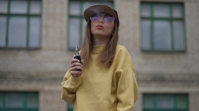 Pretty young hipster woman in black cap yellow hoodie vape ecig. Girl vape popular ecig gadget, vaping device.Happy brunette vaper girl with e-cig. Portrait of smoker female model with electronic