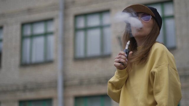 Pretty young hipster woman in black cap yellow hoodie vape ecig. Girl vape popular ecig gadget, vaping device.Happy brunette vaper girl with e-cig. Portrait of smoker female model with electronic