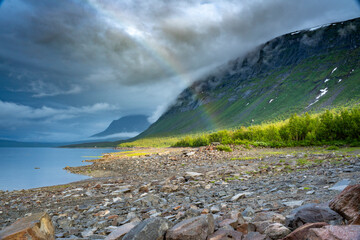 Great View of Cloudy Weather and sunlight with Rainbow breaking through next to Calm Mirror lake Akkajaure in Big Lake National Park In Lapland, northern Sweden. Idyllic and Picturesque Evening Scene.