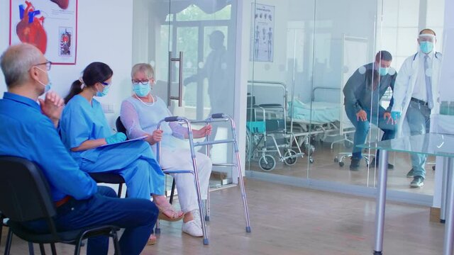 Nurse in hospital waiting room with face mask against covid-19 helping invalid senior woman to fill documents. Doctor inviting old man in examination room. Health care system during global pandemic