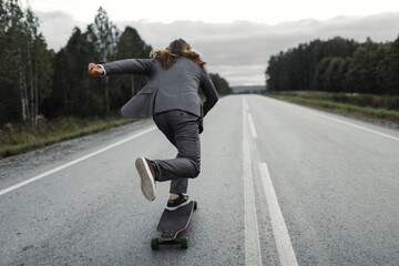 Man in grey office suit with long blond hairs is riding skateboard longboard down road outside the city, back view. Freedom from office work concept. He is riding hands up and enjoying his trip.
