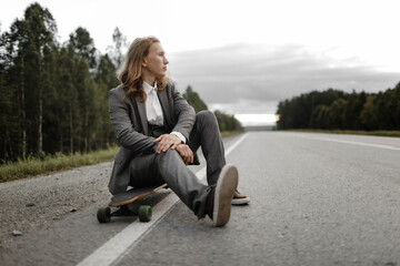 Fototapeta na wymiar Man with long blond hairs in grey office suit with skateboard longboard in city outskirts. He is going to ride a skateboard, front view. Freedom from office work concept.