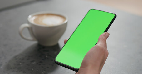 young woman holding smartphone with green screen and drinking cappuccino