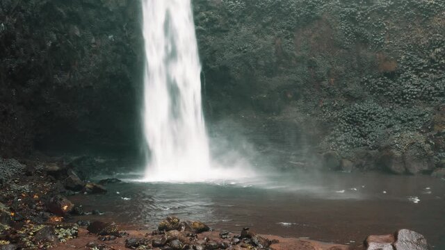 Amazing Nungnung waterfall, Falling water hitting water surface, some huge rocks seeable in front of frame. Lush green leafes is moving from the wind, Bali, Indonesia