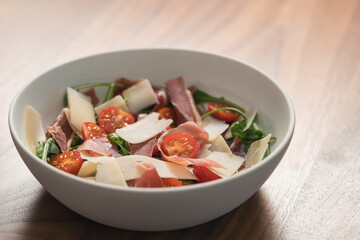 Salad with prosciutto, tomato, arugula leaves and parmesan cheese in white bowl on walnut table