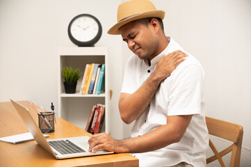 Young asian man in white shirt with hat sitting in home working with laptop he has shoulder, neck pain and hurt get stress he symptom office syndrome from he working for long time.