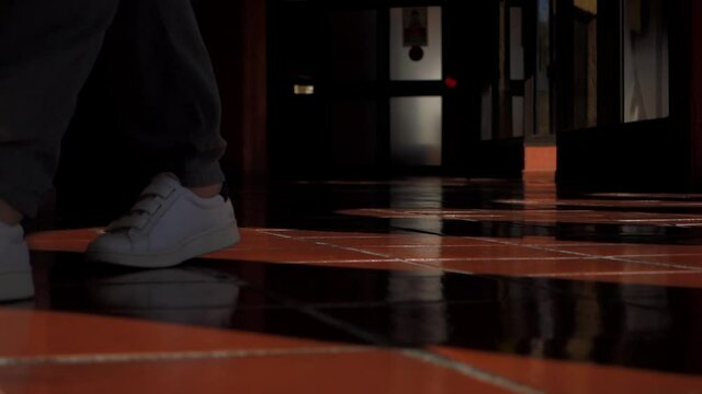 Female feet in sneakers wearing romper go to the camera inside building with light and shadow, low angle filming. Lifestyle concept.