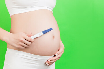 Cropped image of pregnant woman in white underwear showing positive pregnancy test against her abdomen at green background. Childbirth expecting. Copy space
