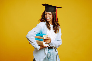 Graduate girl in a graduation hat on her head, with books stands on a yellow background. African American girl posing with smile. Graduation, university, college, distance education concept.