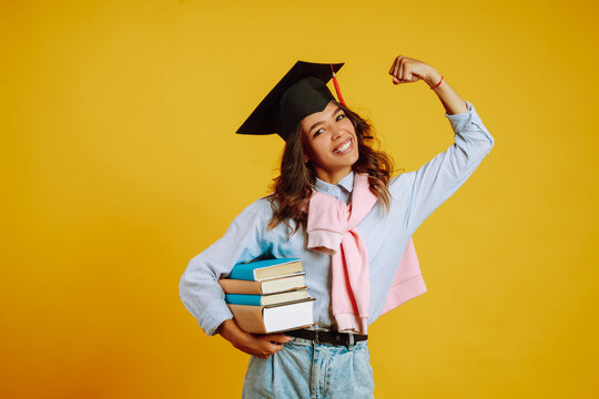 Graduate girl in a graduation hat on her head, with books stands on a yellow background. African American girl posing with smile. Graduation, university, college, distance education concept.