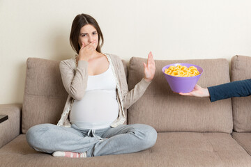 Pregnant woman sitting on the sofa refuses to eat unhealthy snacks. Stop to the junk food during...