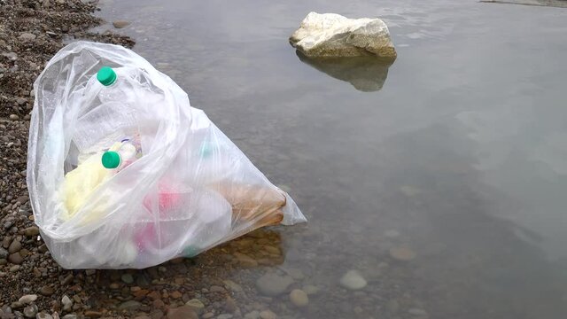 Plastic bag with collected plastic waste lies on the river bank, environmental pollution