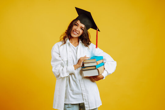 Graduate girl in a graduation hat on her head, with books stands on a yellow background. African American girl posing with smile. Graduation, university, college, distance education concept. - Powered by Adobe