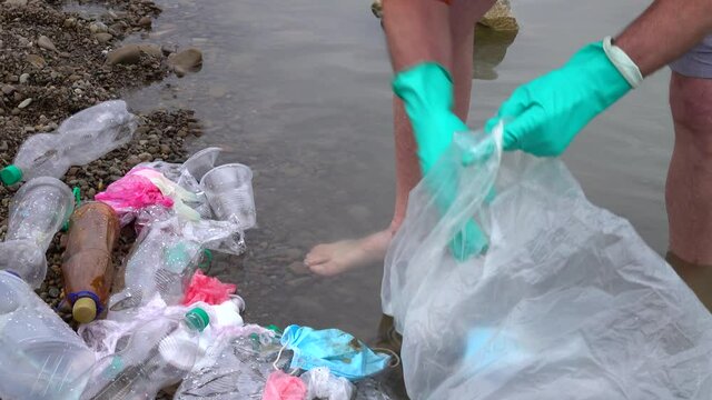 Man in the rubber gloves, standing in the water of the river, picks up plastic waste into a plastic bag