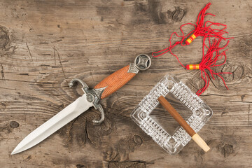 Cigar in ashtray and dagger on old wooden board. Top view.