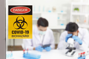Caution symbol and biohazard symbol to stick on glass in front of lab. Two Male Scientists wear...