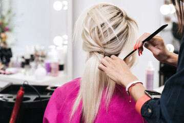 Hands of a hair stylist with a comb make a hairstyle for a blonde girl