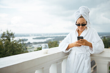 Smiling cute girl blogger in white robe, towel and sunglasses stands on balcony with phone and cup...