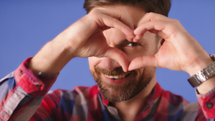Portrait of smiling young man keeps hands over his eye in heart shape , expresses sympathy. Close up. High quality photo