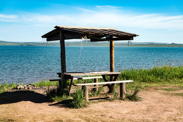 Wooden gazebo with a roof and benches on the shore of a turquoise lake.