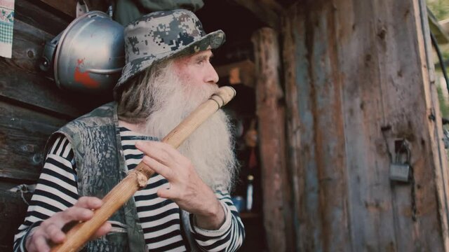 Old hermit grandfather with a gray beard plays a flute he made himself, next to a stringed folk instrument. An old man with dreadlocks looks into the distance, enjoying his creativity.