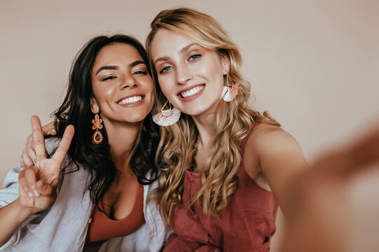 Pretty latin girl with dark hair posing with peace sign. Indoor photo of enthusiastic white woman making selfie with friend.