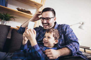 Young father embraces little son family sitting on couch at home using smart phone feels happy...