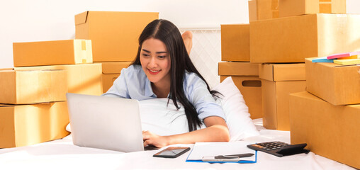 Young smiling beautiful owner asian woman freelancer sme business online shopping working on laptop computer with parcel box on bed at home - SME business online and delivery concept