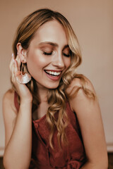 Fototapeta premium Pleasant caucasian woman with long wavy hair laughing with eyes closed. Fascinating stylish lady in earrings standing on light background.
