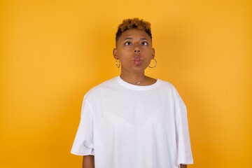 Young African American girl with afro short hair wearing white tshirt standing over isolated yellow wall making fish face with lips, crazy and comical gesture. Funny expression.