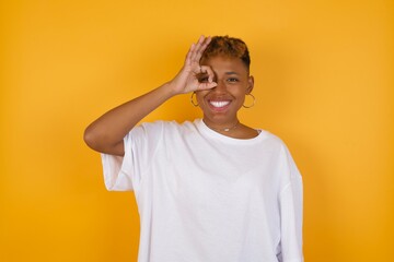 Young African American girl with afro short hair wearing white tshirt standing over isolated yellow wall doing ok gesture with hand smiling, eye looking through fingers with happy face.