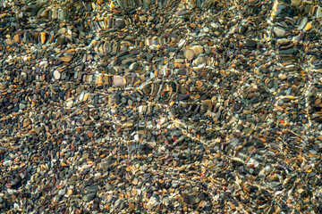 The texture of the bottom. Many small stones are visible under the clear water. Water ripples of the sea, sun rays on the rocks.