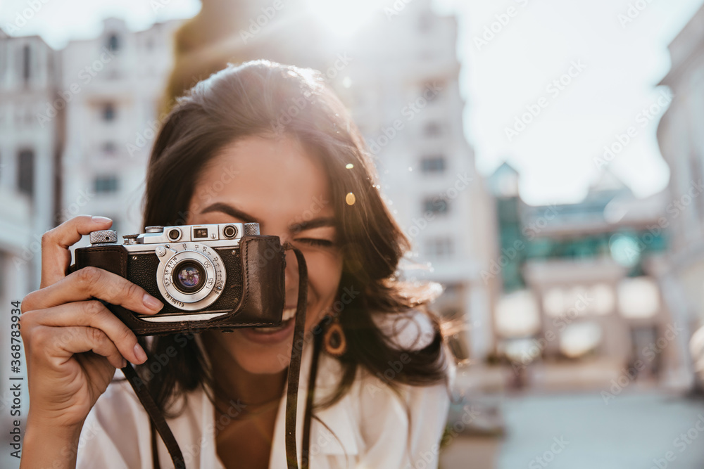 Wall mural Inspired female photographer with dark hair working outdoor. Portrait of smiling girl with camera making photos on the street.