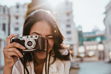 Inspired female photographer with dark hair working outdoor. Portrait of smiling girl with camera making photos on the street. - obrazy, fototapety, plakaty