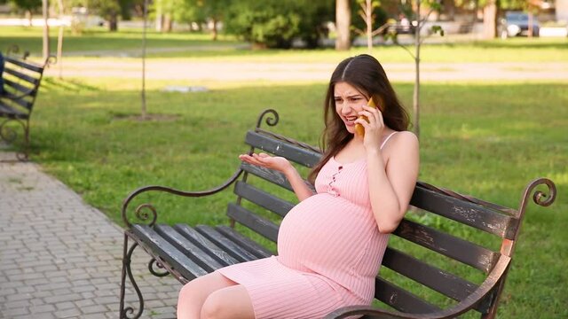 angry pregnant woman sitting on a bench in a summer city park and talking and cursing on her mobile phone