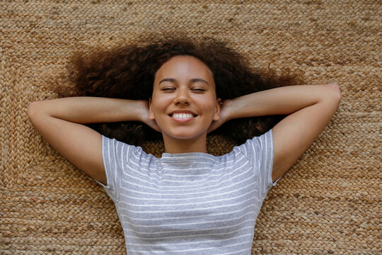 Top view portrait of young woman with dark skin and long curly hair lying on the floor. African american female on wicker rug. Freelancer at home office. Close up, copy space, background.