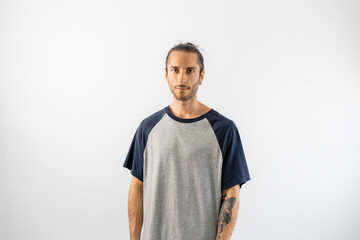 Fototapeta premium Hombre con pelo recogido, camiseta gris de mangas azules y tatuajes posa con rostro tranquilo en fondo blanco