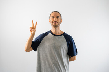Fototapeta premium Hombre con moño y camiseta gris de mangas azules hace el gesto de V de victoria, o número 2, en un fondo blanco aislado