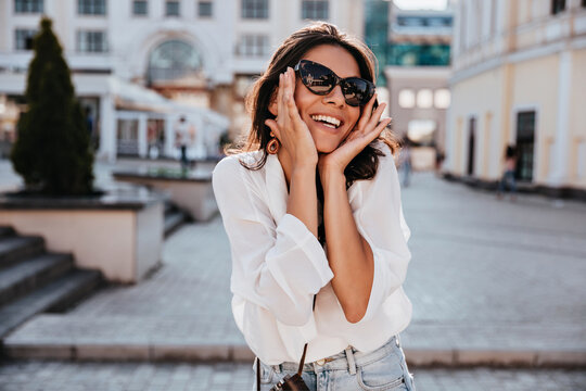 Stunning tanned girl in white blouse enjoying spring weekend. Outdoor photo of good-looking caucasian woman in black sunglasses laughing on the street.