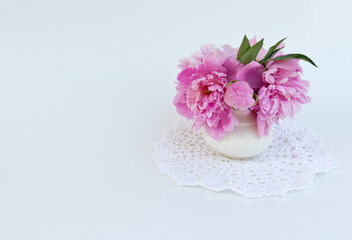 Obraz na płótnie Canvas A bouquet of beautiful delicate pink peonies in a vase on a white lace napkin, handmade crochet. White background. Place for text or congratulations, greeting card, holiday message. Still life