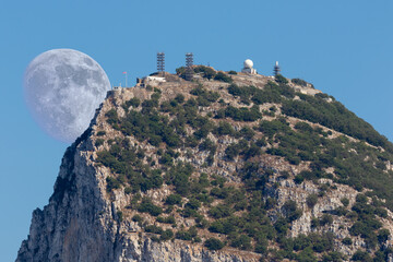 Fototapeta premium Telephoto of the northern summit of Gibraltar with a radio station in the sunshine and blue sky. The moon can be seen big behind the top of the huge rock.
