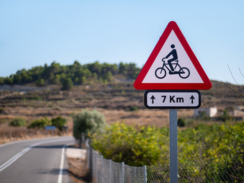 A red and white triangular sign warns of cyclists over a distance of seven kilometers. It is a country road in summer in Spain. The mountains are out of focus in the background.